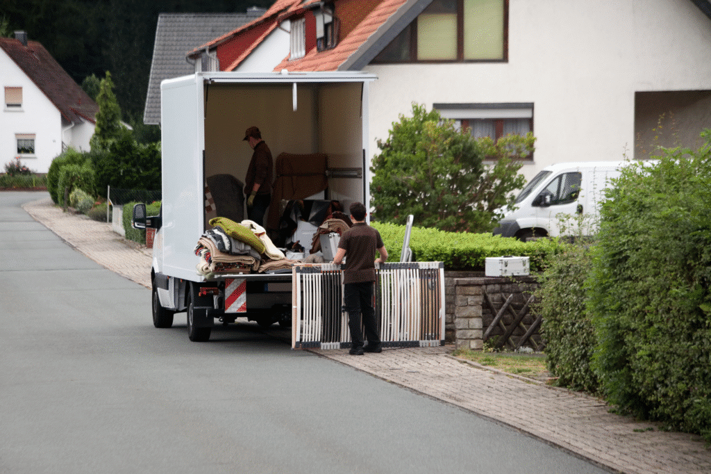 Zu sehen ist ein Transporter. Zwei Männer beladen das Kraftfahrzeug mit einem Bett und jede Menge Gerümpel.