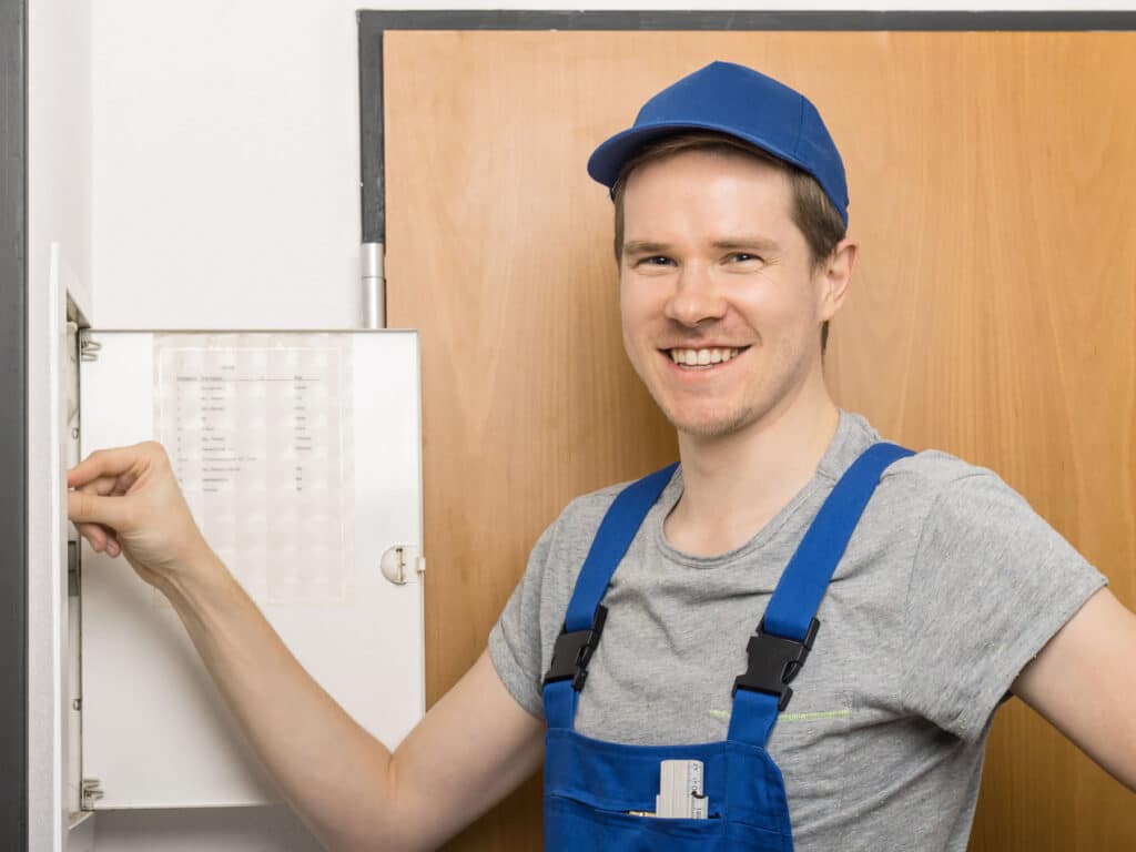 Ein freundlicher Mann, steht vor einen Sicherungskasten und lächelt. Vielleicht sind nur die Sicherungen rausgefallen.